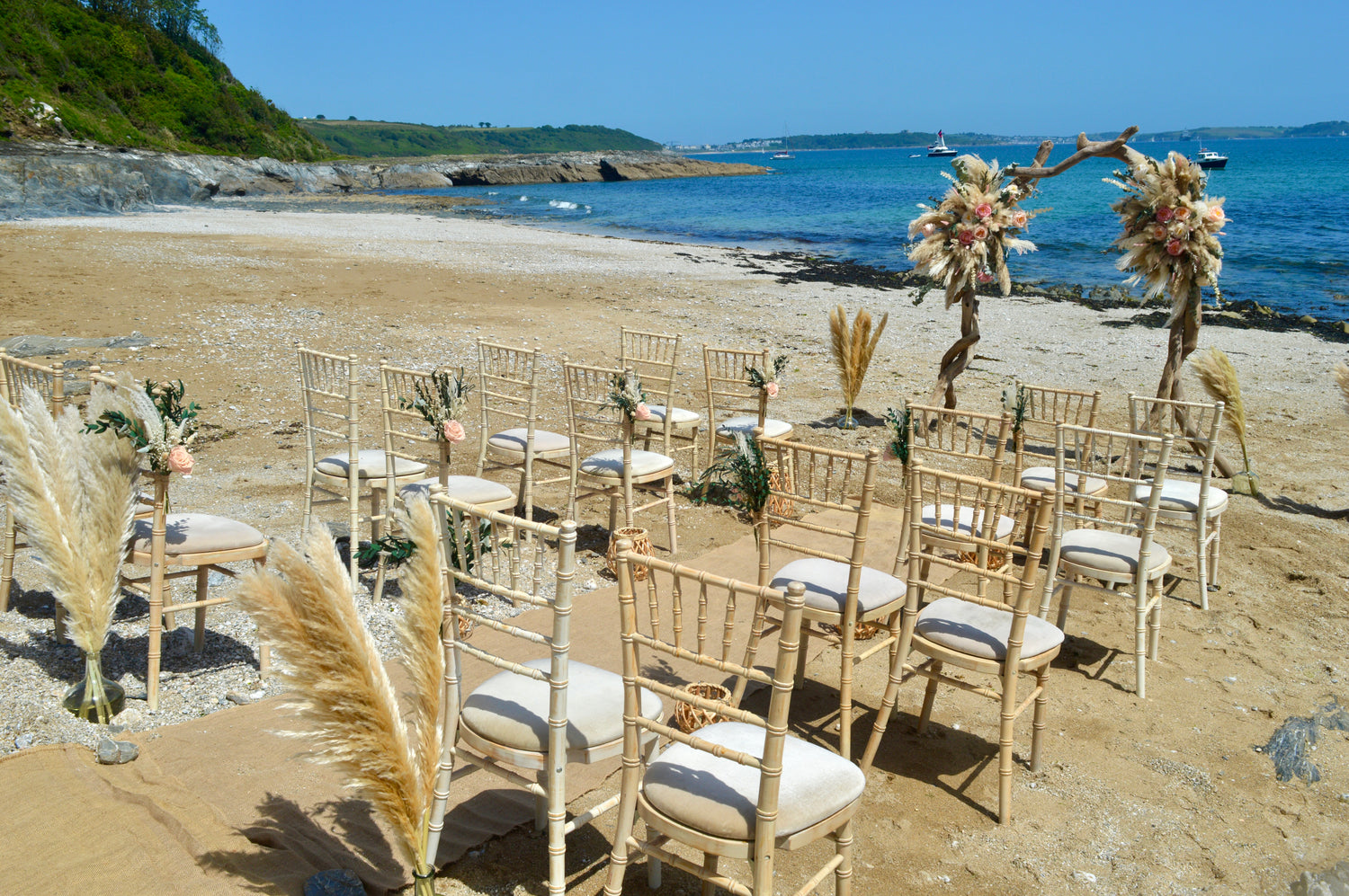 Outdoor beach wedding setup with rows of light wood Chiavari chairs with dried flower decor, cream cushions, pampas grass, floral arrangements, and a driftwood arch with dried flowers on a sandy shoreline with blue ocean and sky.