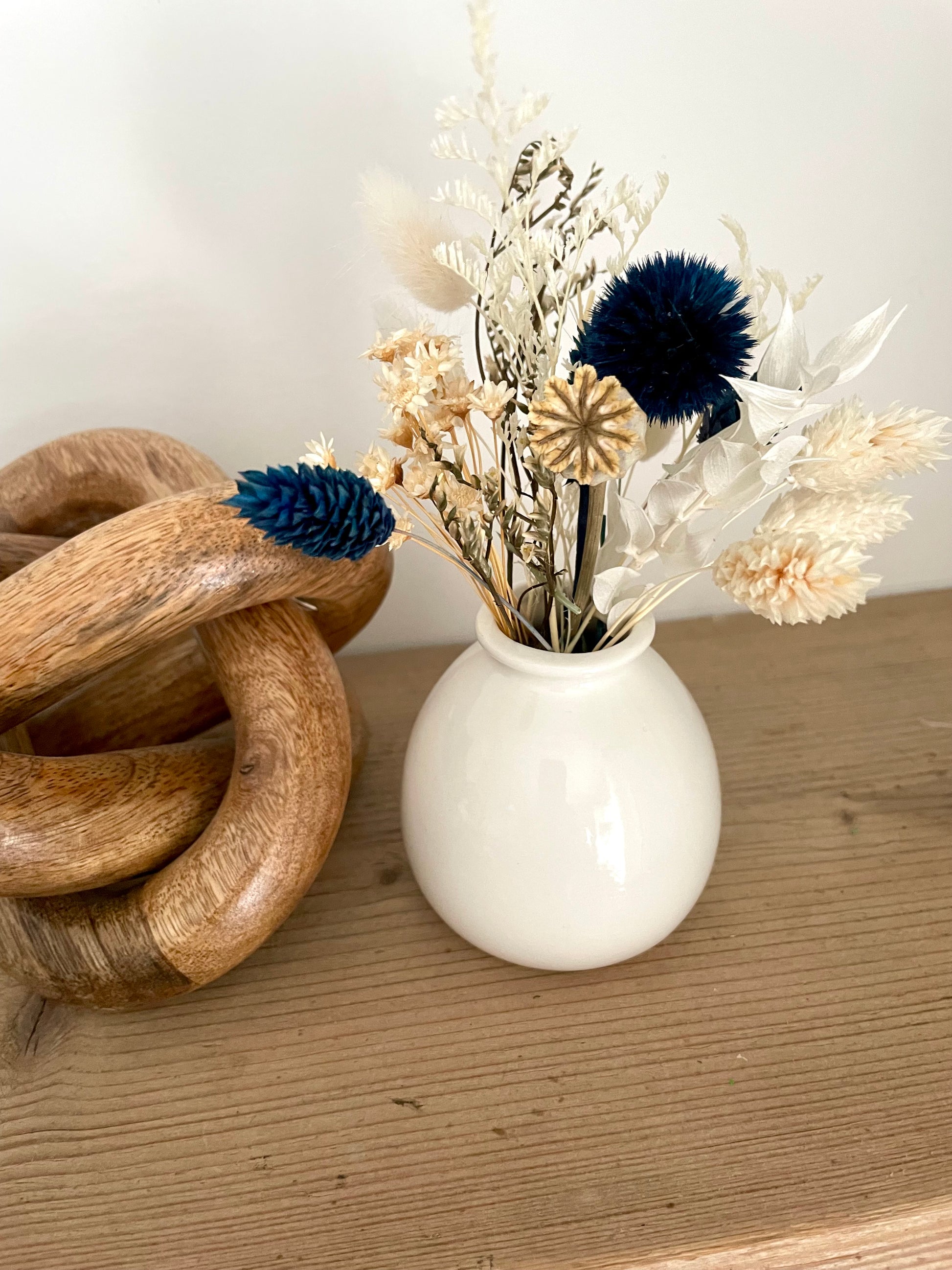 White vase with dried flowers on a wooden surface next to a wooden knot